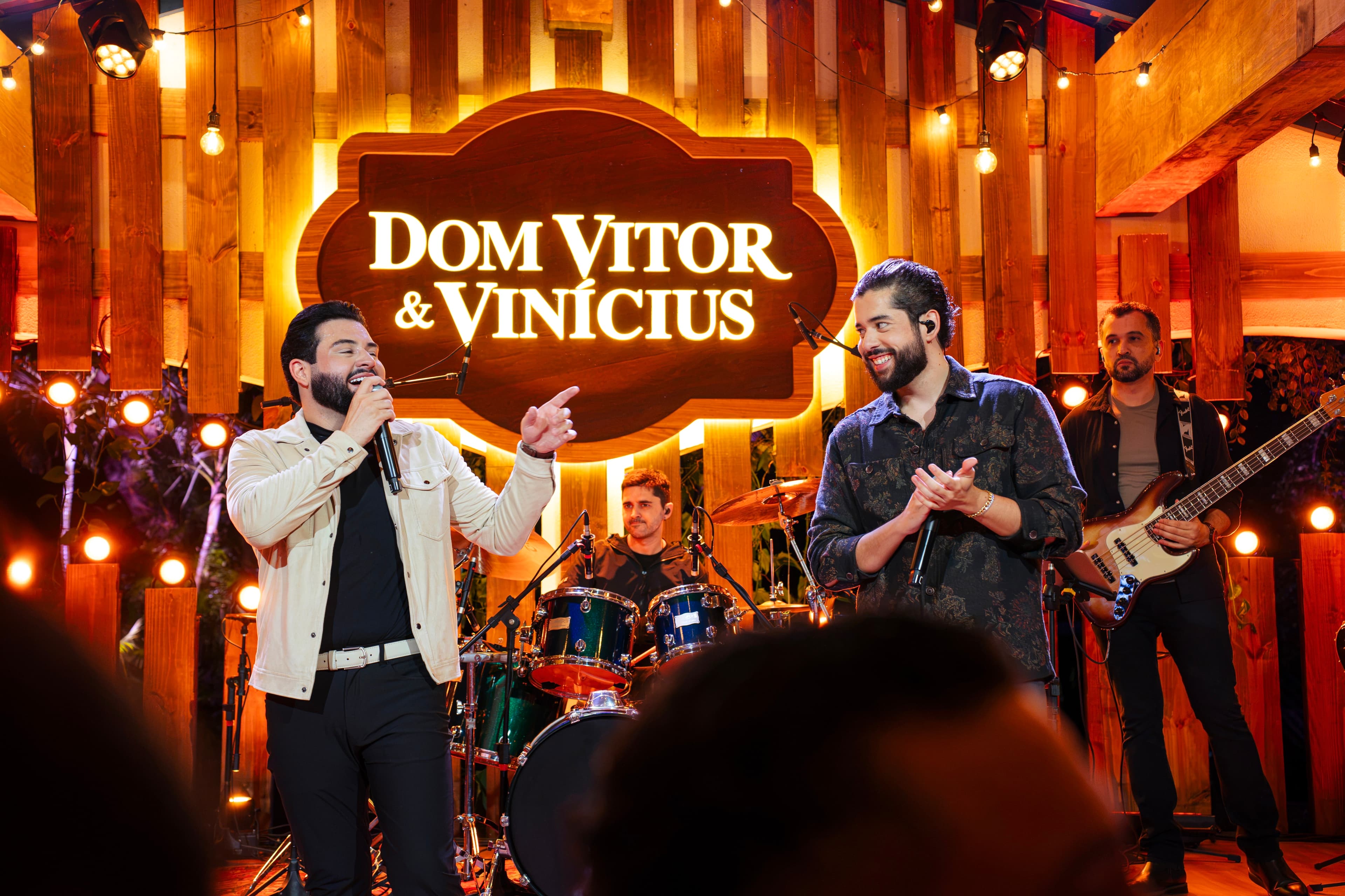 Dupla Dom Vitor e Vinícius posando em frente a um letreiro de LED com o nome da dupla, enquanto, ao fundo, aparecem os músicos da banda.